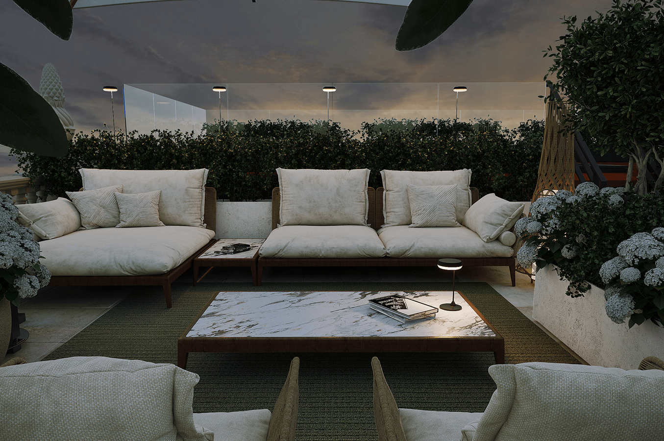 Outdoor seating area at dusk with beige cushioned sofas and armchairs around a marble coffee table, surrounded by green plants and white flowers.